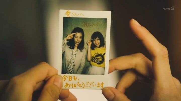 two hands hold a photograph of a 29  year old woman and a younger idol wearing a yellow "sunny side up" shirt